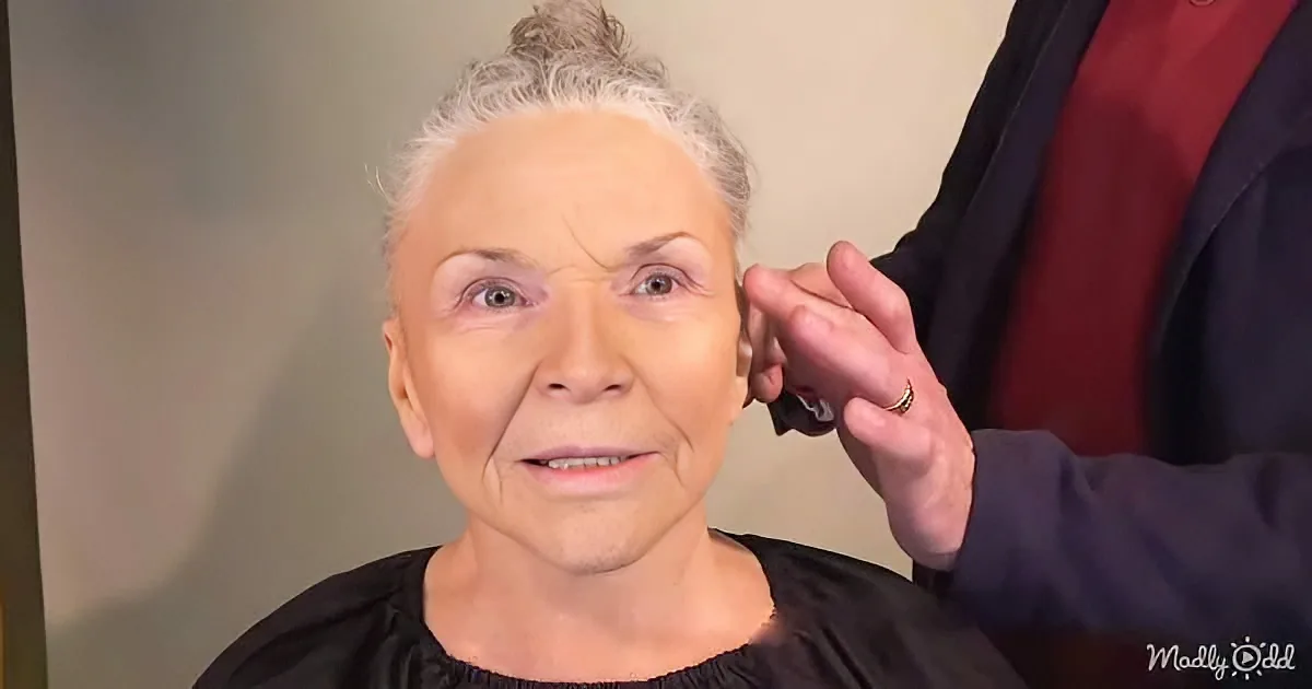 An older woman with gray hair in a bun, wearing a black top, is having her ear examined by someone in a red and black outfit.