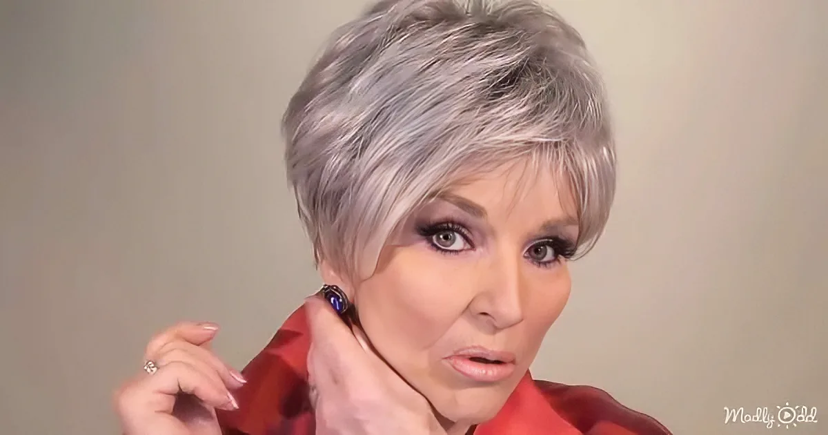 A woman with short, gray hair and a red garment adjusts her earring.