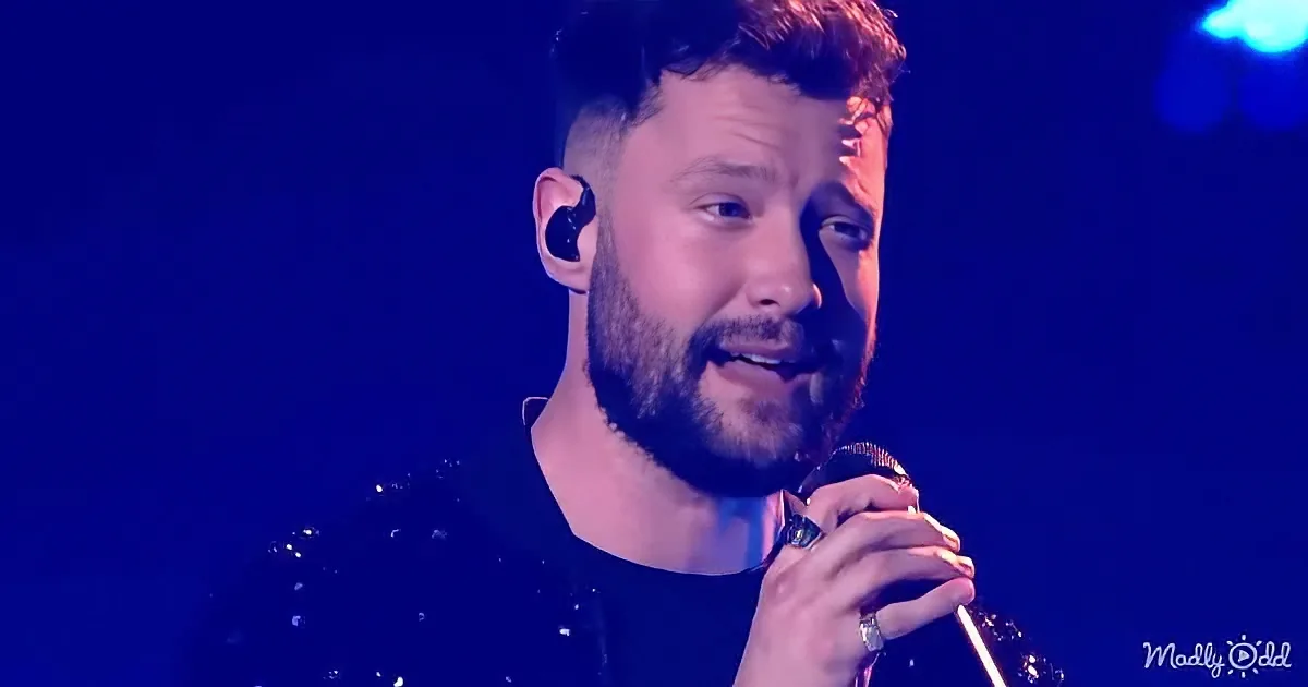 A man with a beard and earpiece sings into a microphone under blue stage lighting.