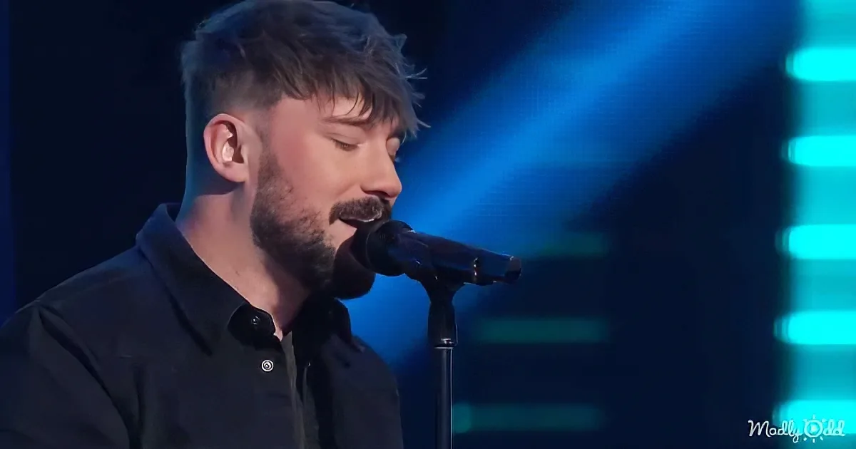 A man with a beard sings into a microphone on stage, illuminated by blue lights.