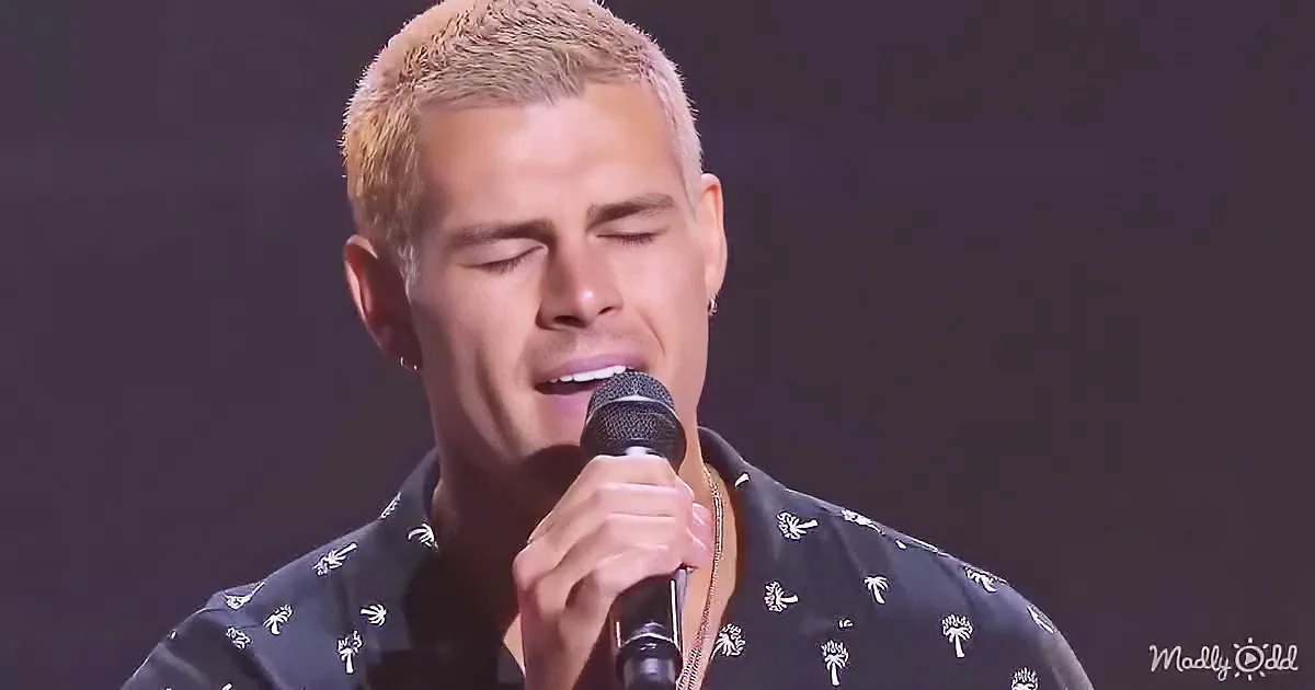A person with short, light-colored hair sings into a microphone, wearing a patterned shirt.