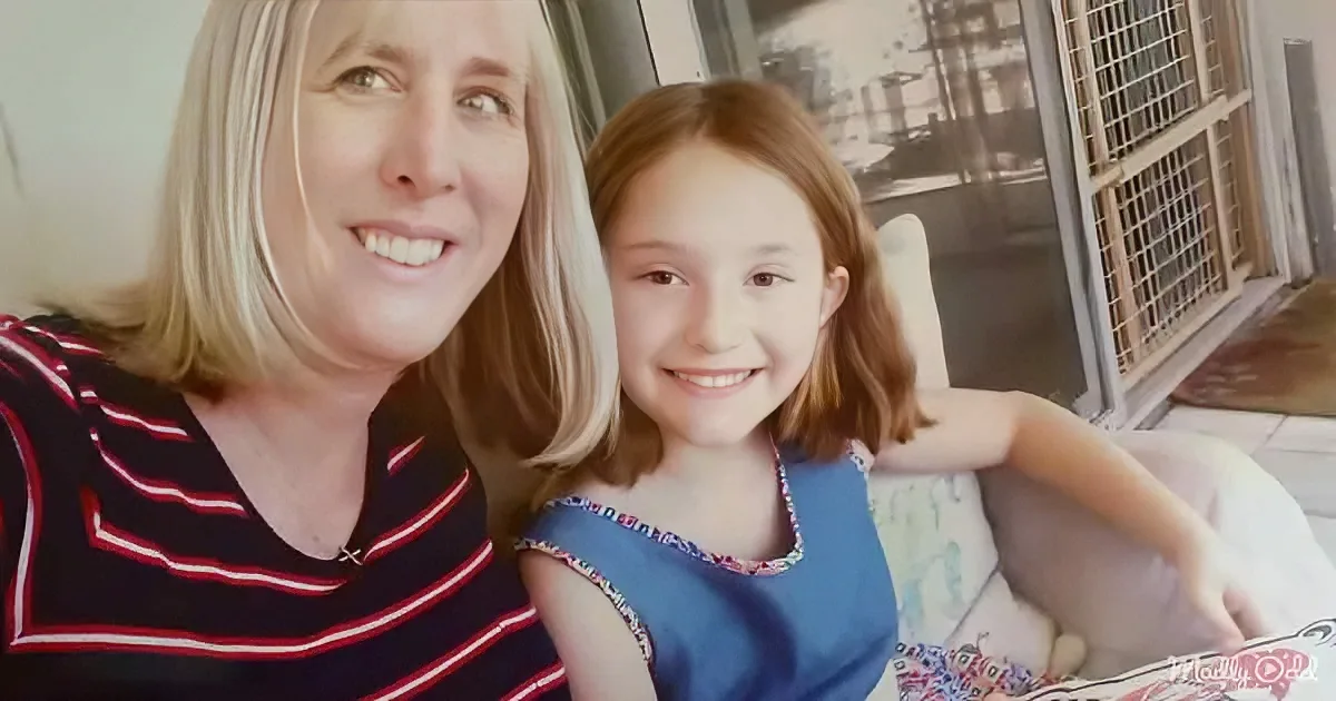 A woman and a young girl smile together, the woman in a striped shirt, the girl in a blue dress, with a window and metal...