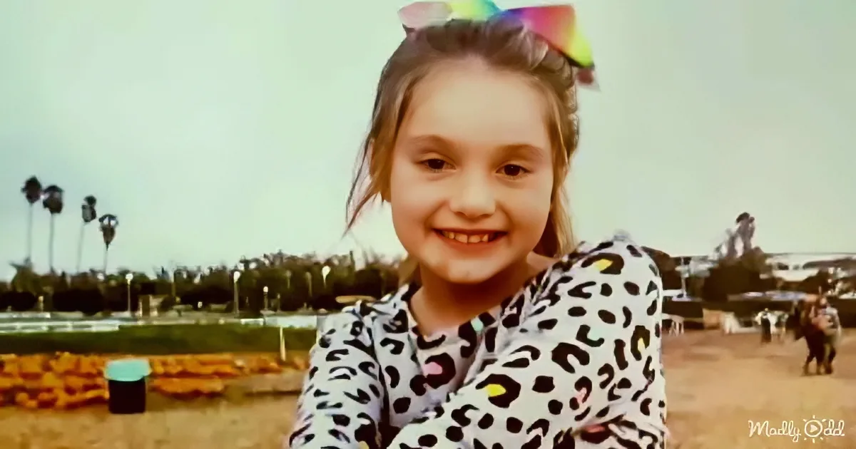 A young girl with a rainbow bow in her hair smiles in a leopard print shirt, with palm trees and a park in the background.