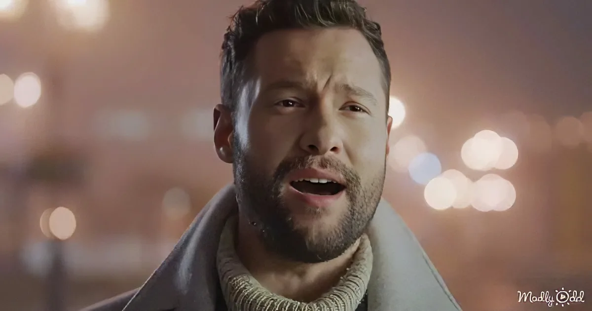 A man with a beard and short hair, wearing a coat and sweater, stands against a blurred background of lights.
