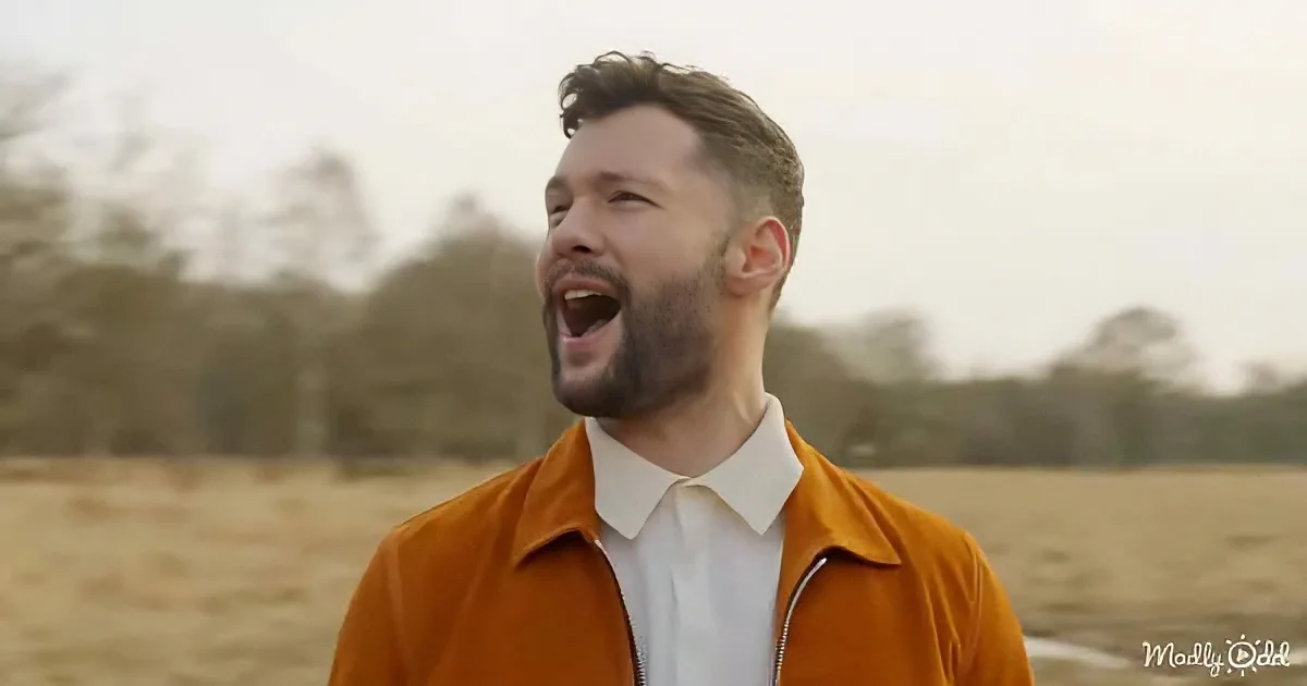 A man in an orange jacket and white shirt stands in a field, mouth open, possibly singing or speaking.