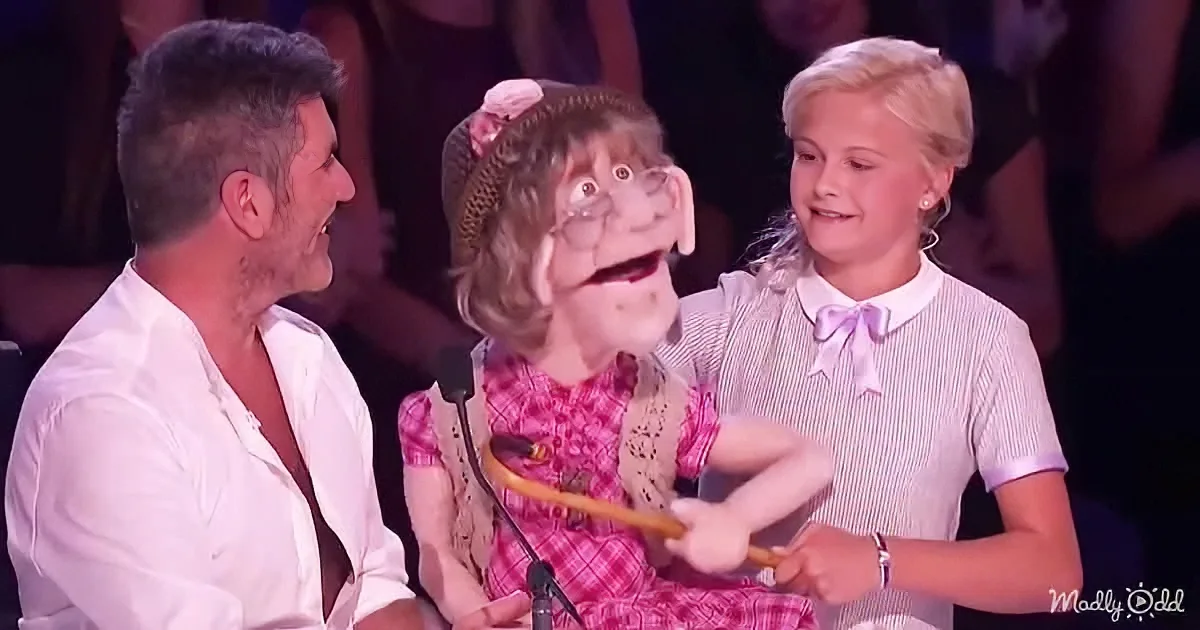 A puppet in a pink dress holds a stick, surrounded by two people, with a crowd in the background.