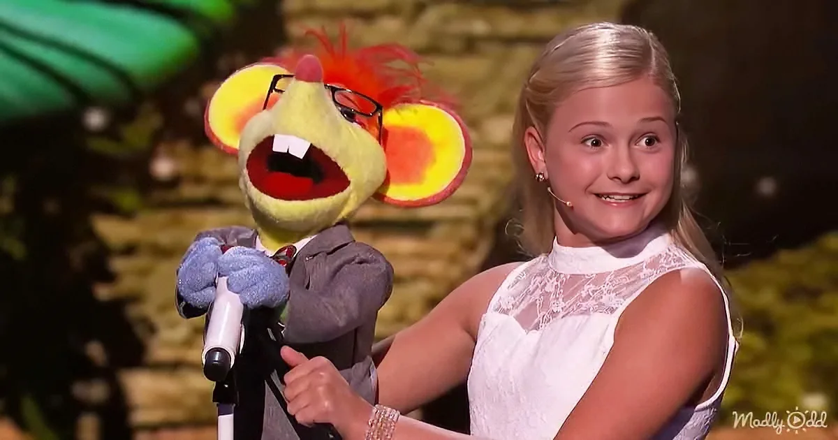 A young girl in a white dress stands next to a puppet with a microphone, both appearing surprised.