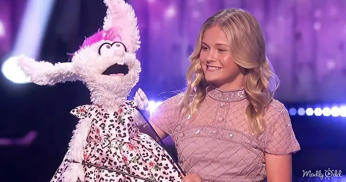 A young woman in a pink dress holds a white puppet with a pink hat and leopard print dress on stage.
