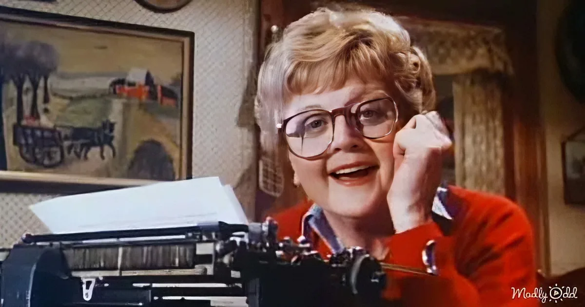 A smiling woman in a red sweater sits at a typewriter, holding her hand to her face.