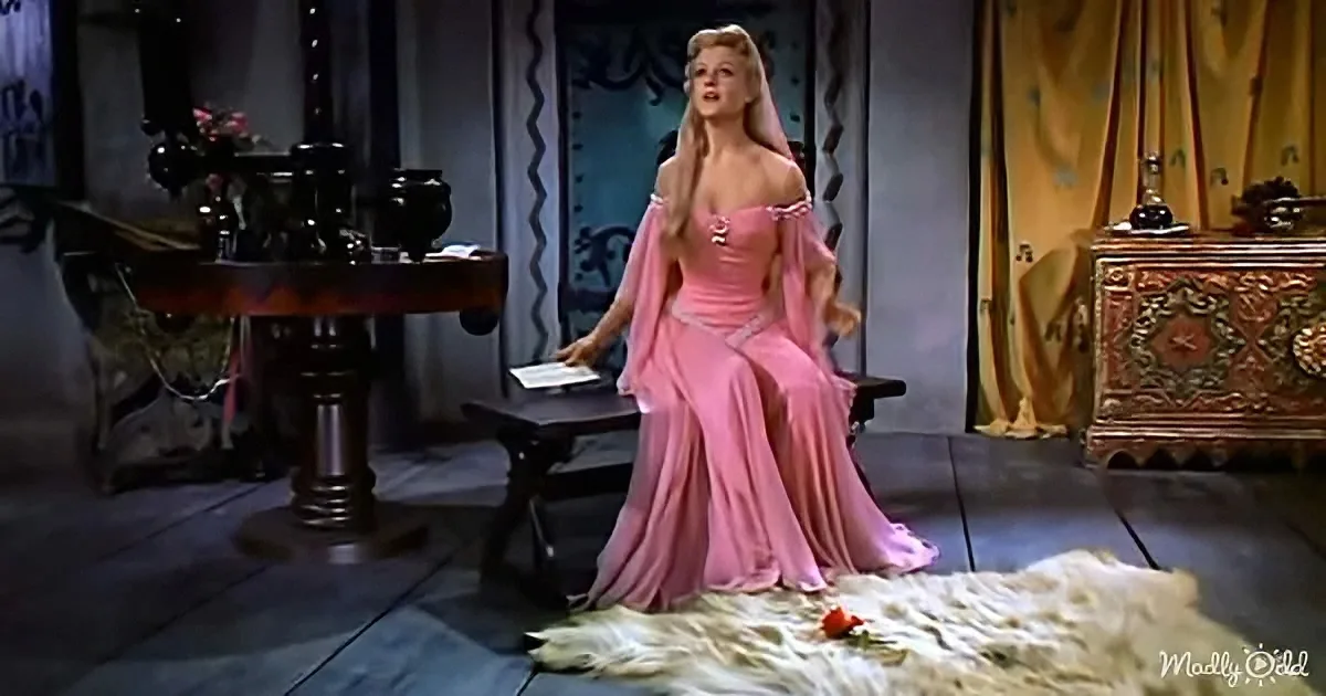 A woman in a pink gown stands in a dimly lit room, holding a book, with a table and ornate furniture in the background.