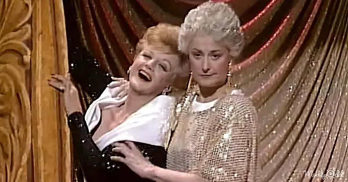 A woman in a white dress is hugging another woman in a glittery outfit, both smiling and laughing.