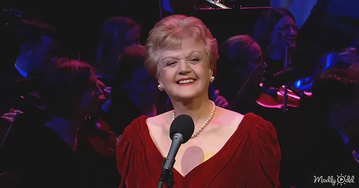 A woman in a red dress smiles at a microphone, surrounded by an orchestra.