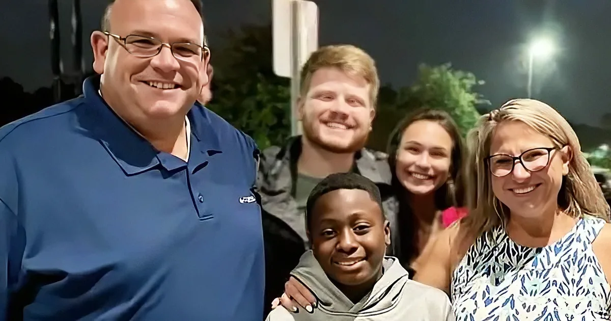 A man in a blue shirt smiles alongside a group of people, including a young boy, under evening lights.