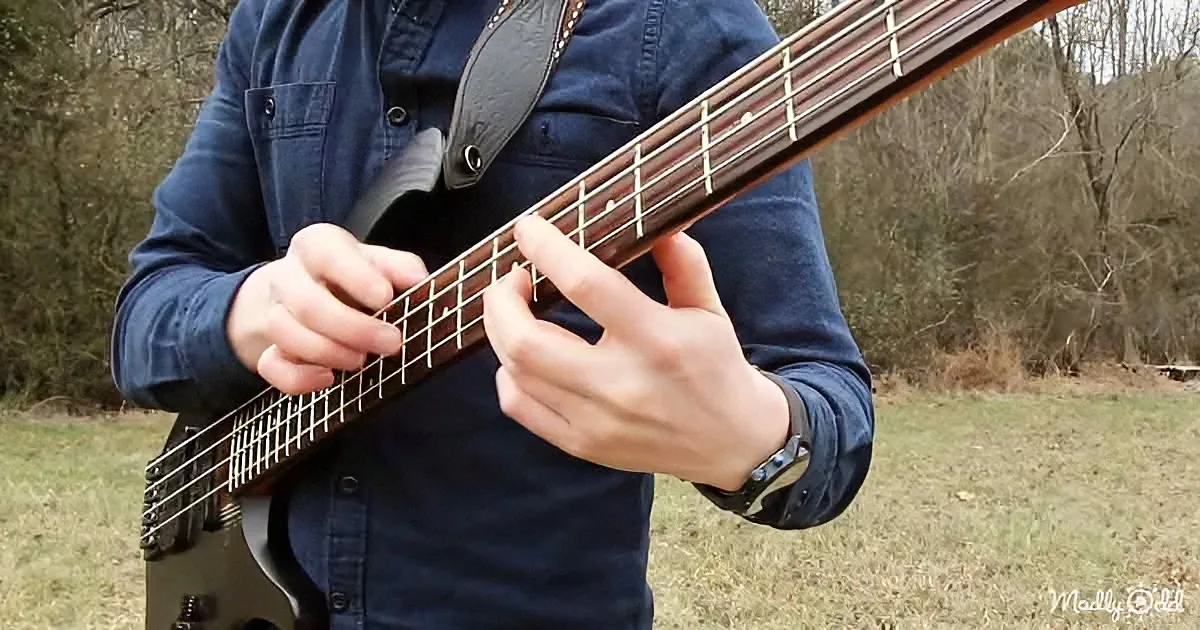 A person plays a bass guitar outdoors, wearing a blue shirt and a watch, with a forested background.