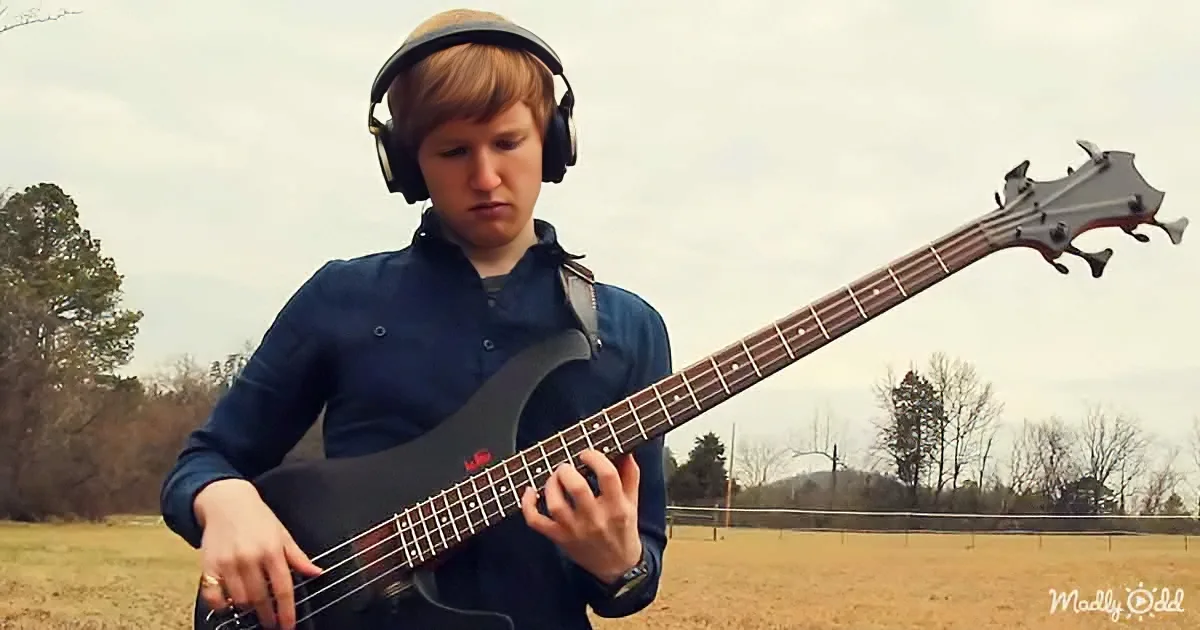 A person wearing headphones plays a bass guitar in an open field with trees in the background.