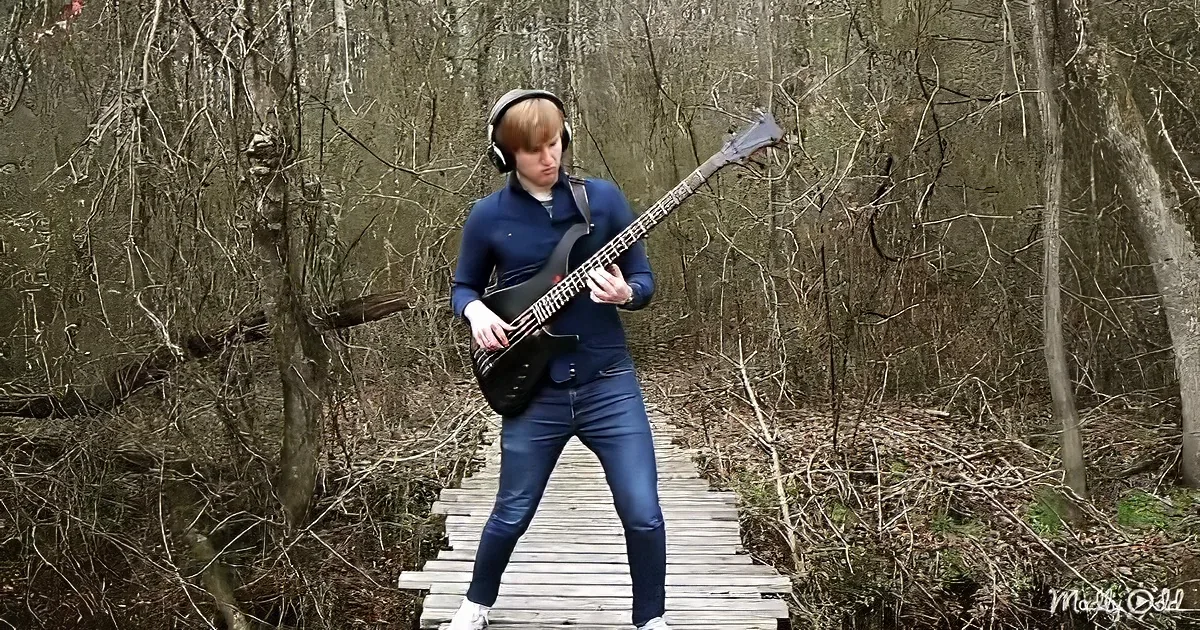 A person wearing headphones plays a bass guitar on a wooden bridge in a forested area.