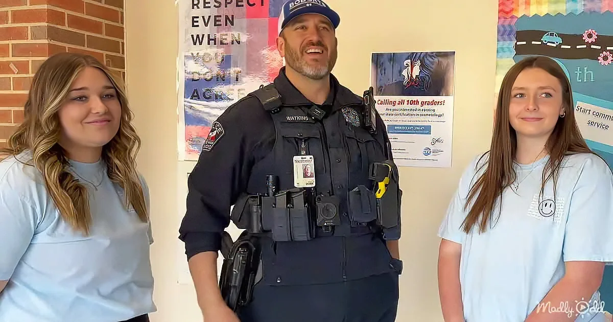 A police officer in uniform stands between two young women in a school setting.