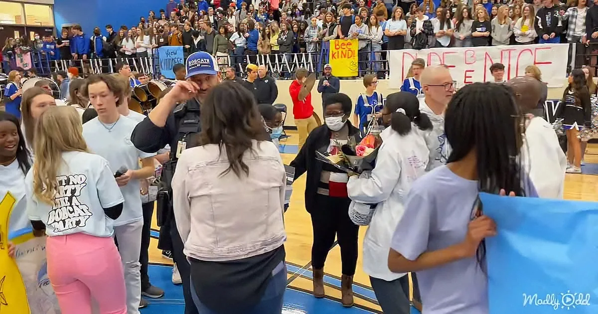 A woman in a black outfit holds flowers, surrounded by students and a police officer in a gymnasium.