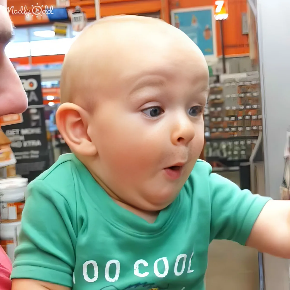 A baby in a green "Too Cool" shirt looks surprised in a store setting.