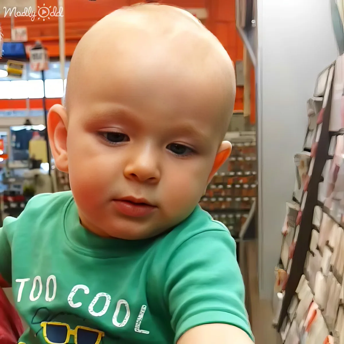 A baby with a shaved head wearing a green "Too Cool" shirt in a store.