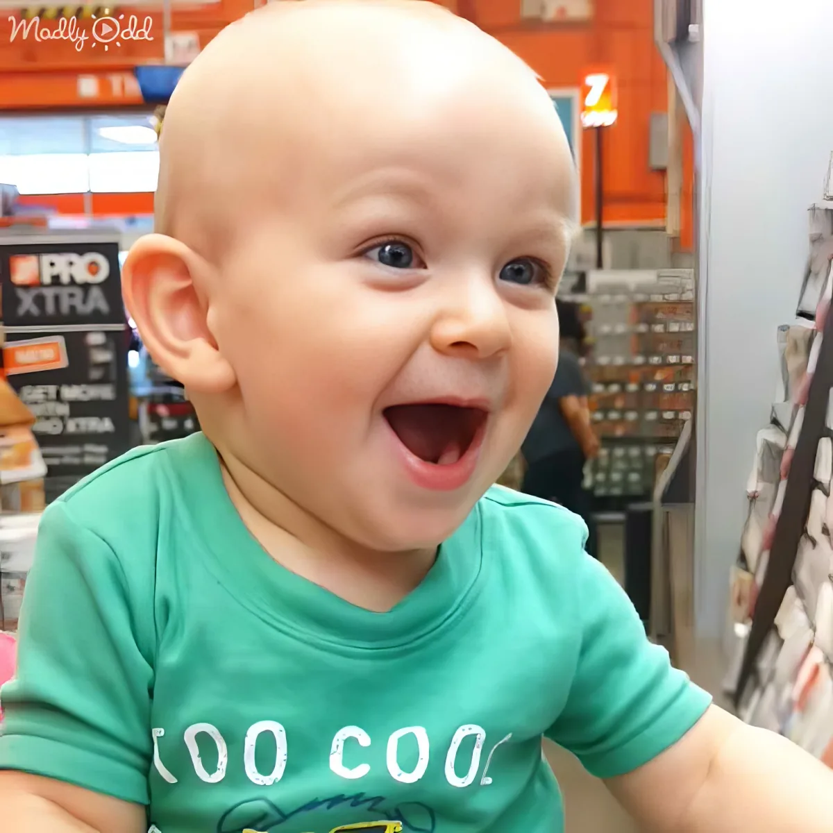 A joyful baby in a green "Too Cool" shirt smiles widely in a store setting.