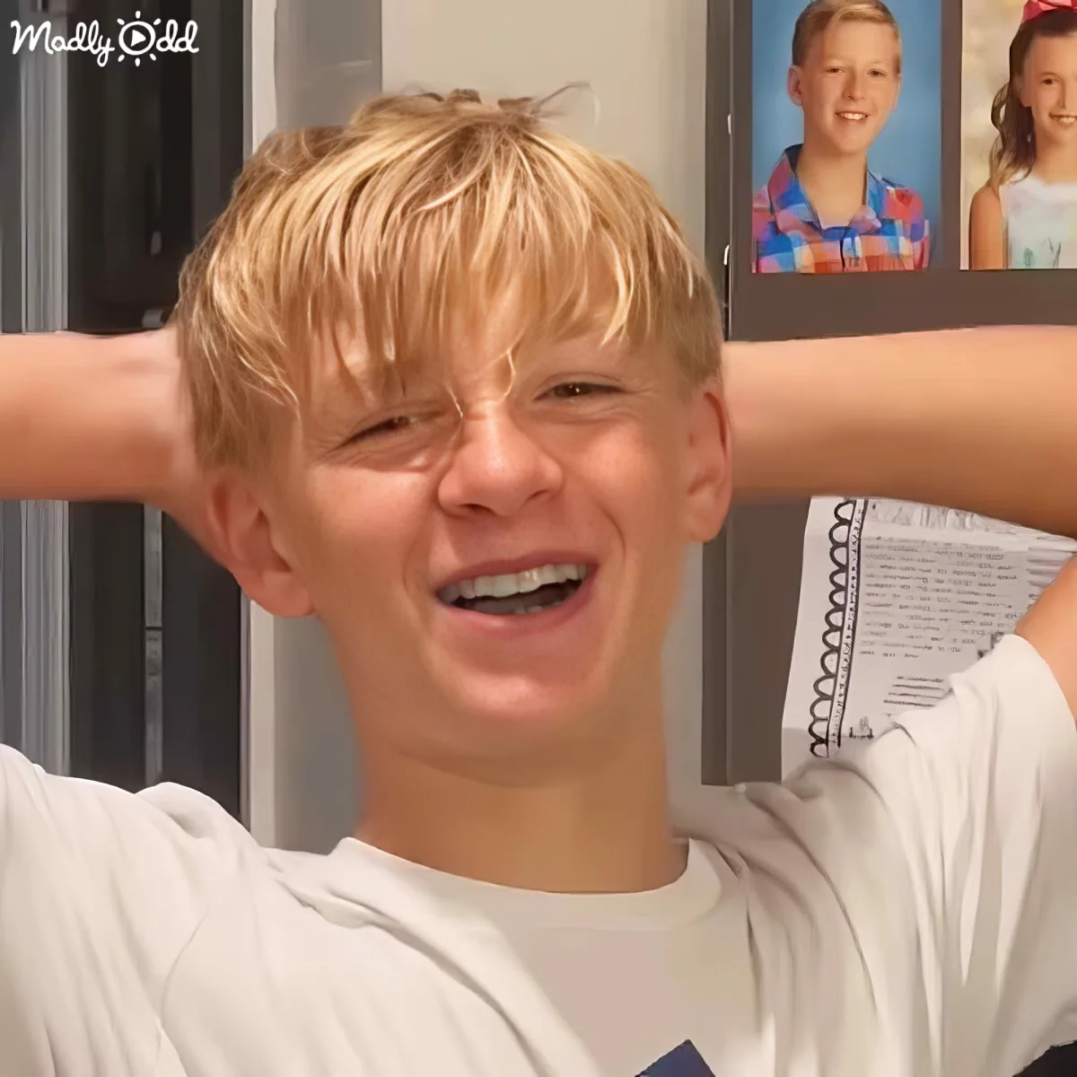 A boy with blonde hair and braces smiles widely, arms outstretched, in front of a photo frame.