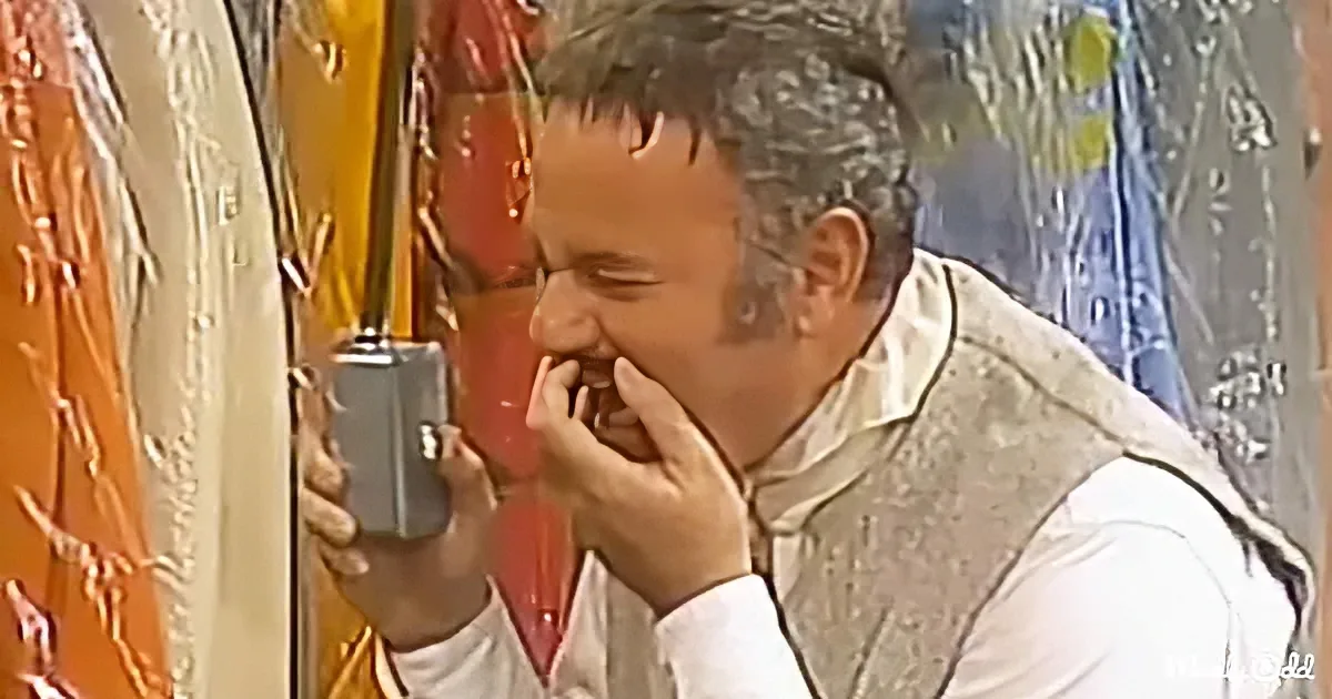 A man in traditional attire holds a phone, appearing to cry or laugh, with a colorful background.