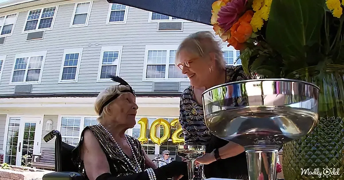 An elderly woman in a black dress and feather headband is being assisted by another woman at a table with a large silver...