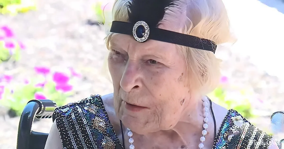 An elderly woman wearing a black sequined dress, pearl necklace, and a black headband with a brooch, sits in a wheelchair...