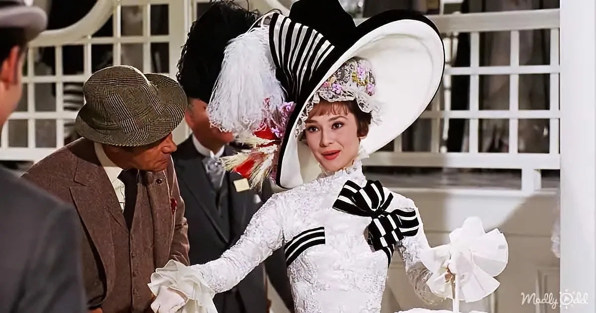 Audrey Hepburn in a white lace dress and large hat, holding a white ribbon, surrounded by men in suits.