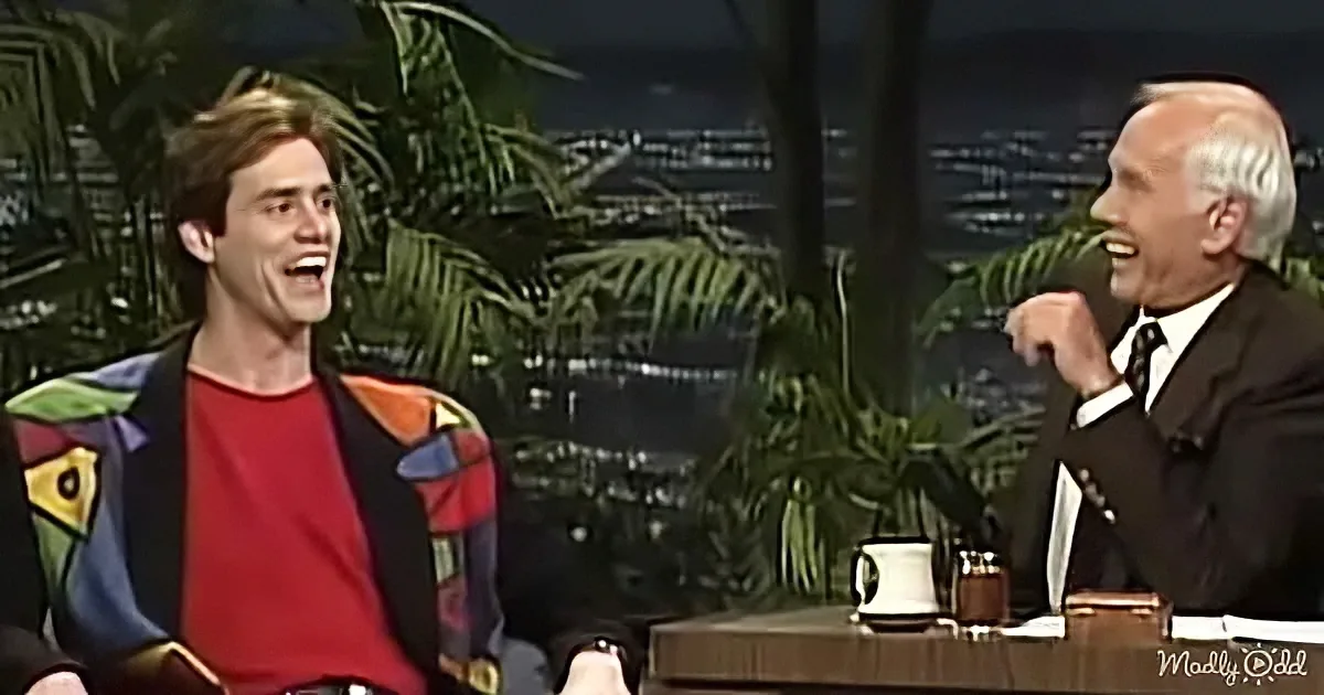 A man in a colorful jacket laughs alongside another man in a suit on a talk show set.