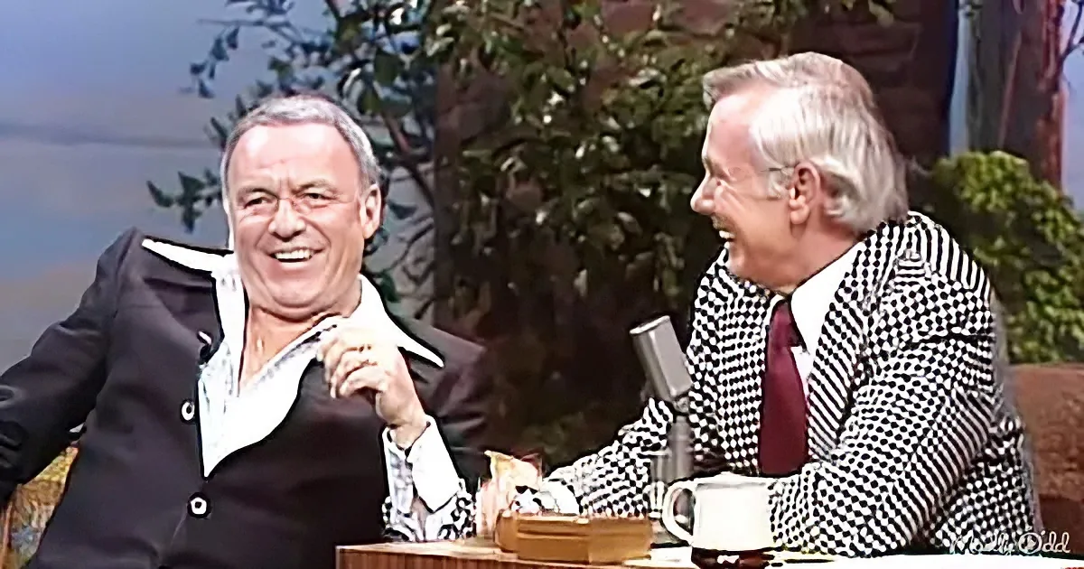 Two men in suits are laughing, one in a black suit and the other in a checkered suit, seated at a table with a microphone...