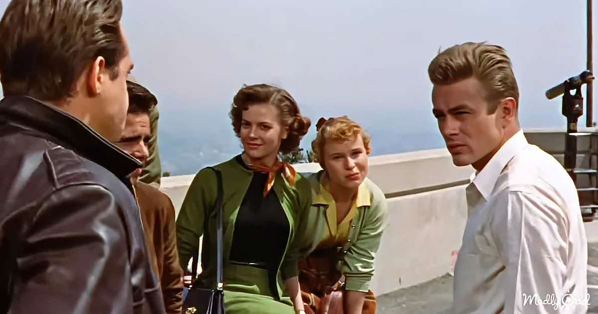 James Dean stands facing a group of people, including a woman in a green dress, in a scene from "Rebel Without a Cause."