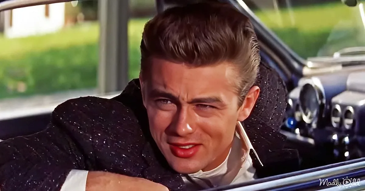 A man with slicked-back hair leans on a car door, wearing a dark jacket and white shirt, with a serious expression.