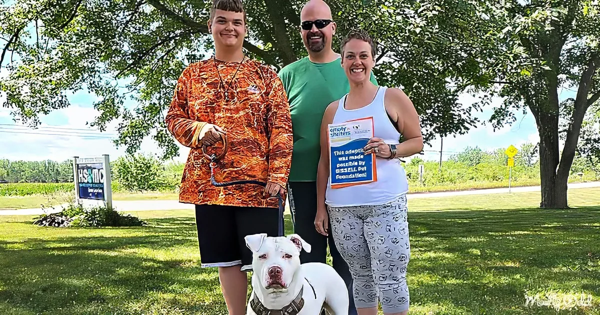 A family stands outdoors with a white dog, holding a sign about pet adoption.