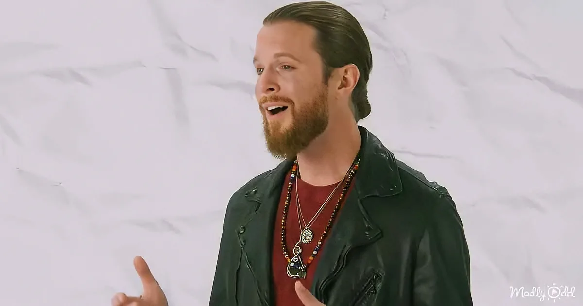 A man with a beard and ponytail, wearing a black jacket and red shirt, gestures with his hands while speaking.