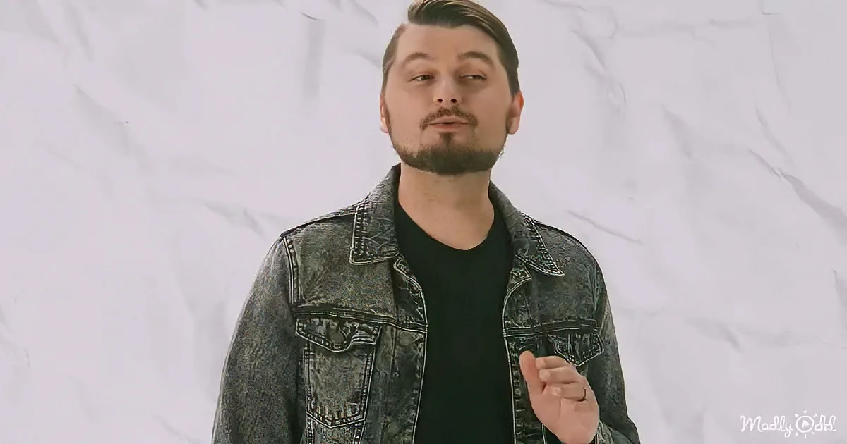 A man with a beard and short hair, wearing a denim jacket over a black shirt, stands against a white, textured background.