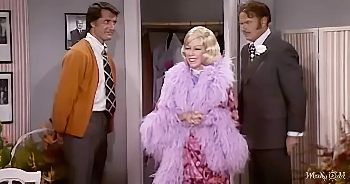 A woman in a purple feathered outfit stands between two men in a room with framed pictures.