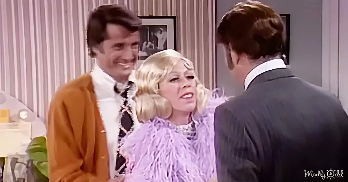 A woman in a purple fringe dress stands between two men, one in a brown cardigan and another in a suit, in a room with...