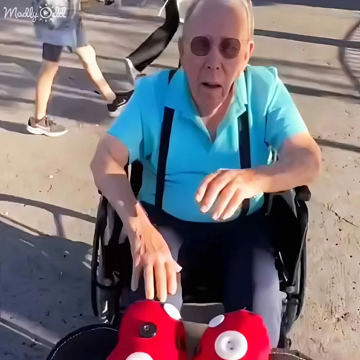 An elderly man in a wheelchair wears a blue shirt and glasses, interacting with red plush toys.