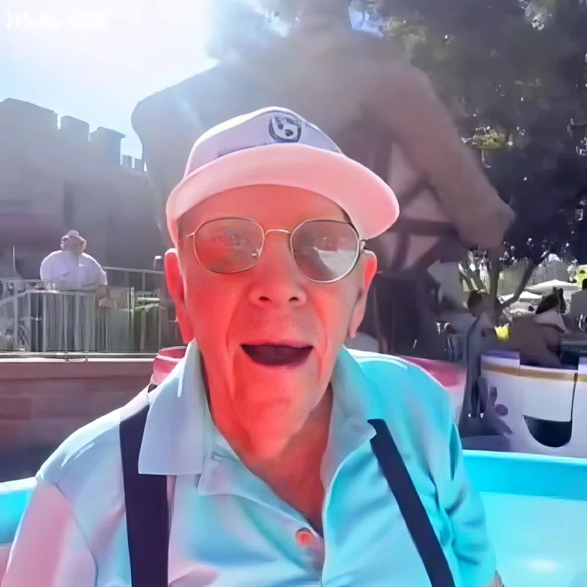 An older man wearing a pink cap, sunglasses, and a light blue shirt is smiling while sitting in a boat at what appears to...