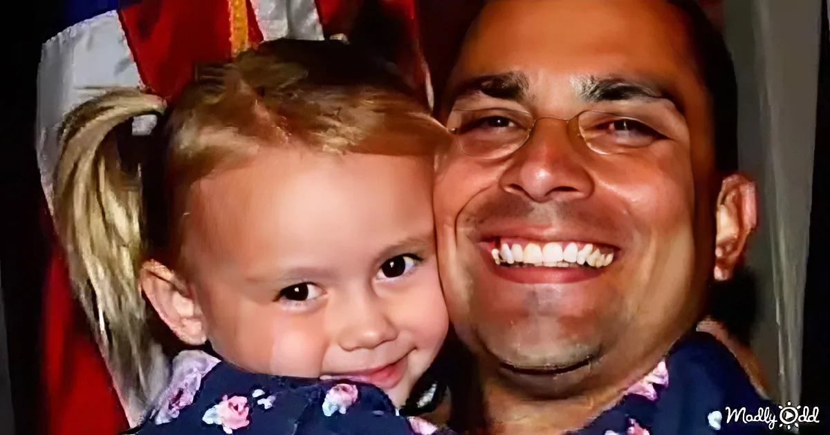 A smiling man holds a young girl with blonde pigtails, both looking at the camera.
