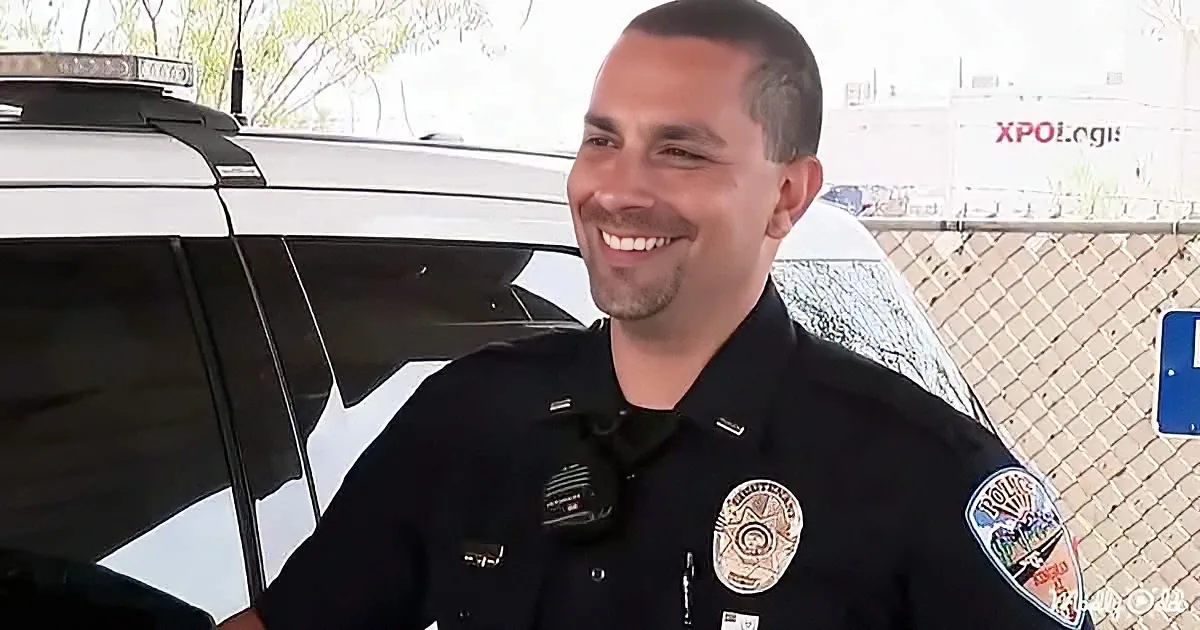 A smiling police officer in uniform stands in front of a police car, with a white truck and chain-link fence in the...