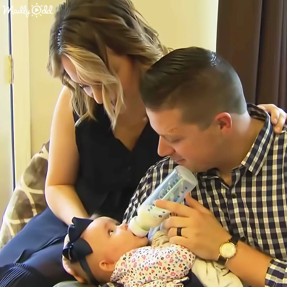 A man feeds a baby with a bottle while a woman watches, both seated indoors.