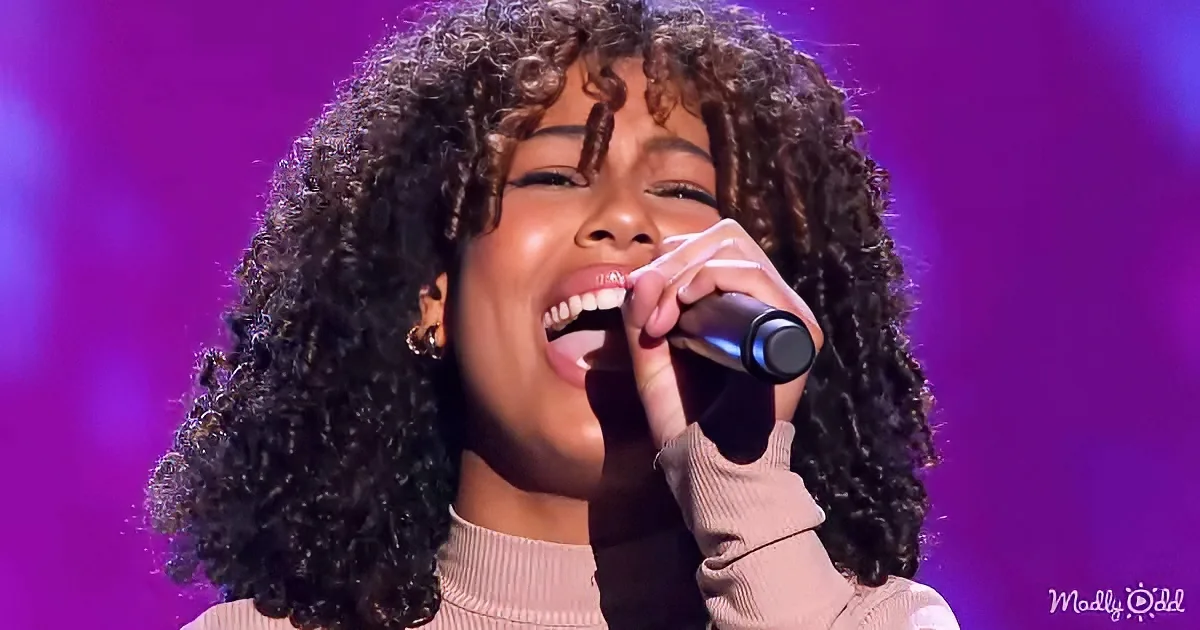 A person with curly hair sings into a microphone against a purple background.