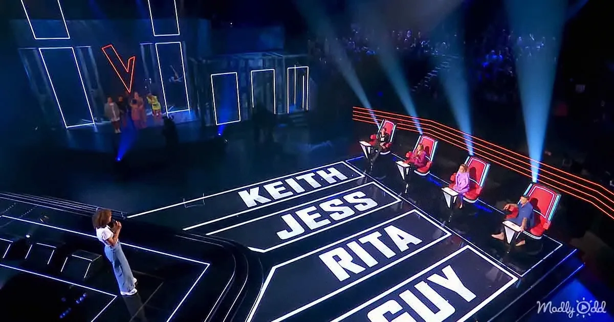 A singer performs on stage, judges sit behind their names, audience in the background.