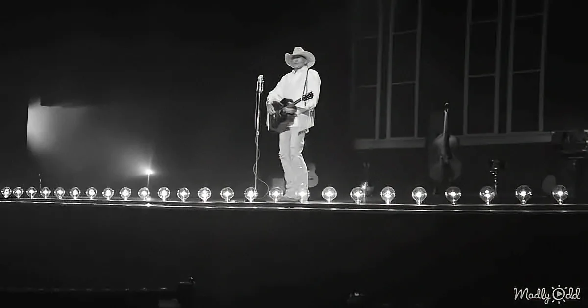 A musician in white attire and a cowboy hat plays guitar on stage with a microphone and string of lights.
