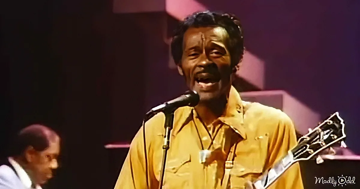 A man in a yellow shirt sings into a microphone while holding a guitar, with another person playing in the background.