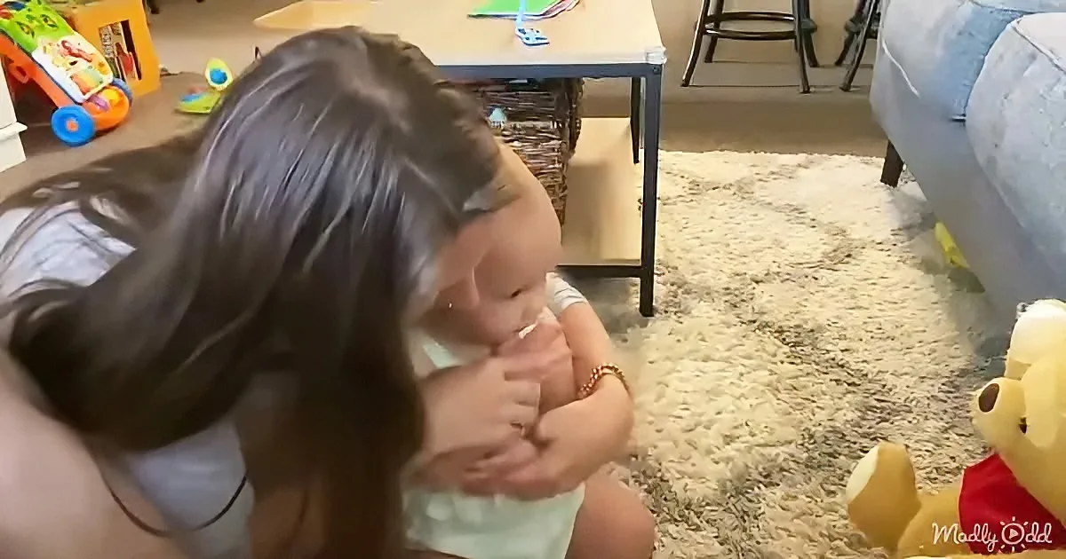 A woman holds a baby on her lap, surrounded by toys and a cozy living room setting.