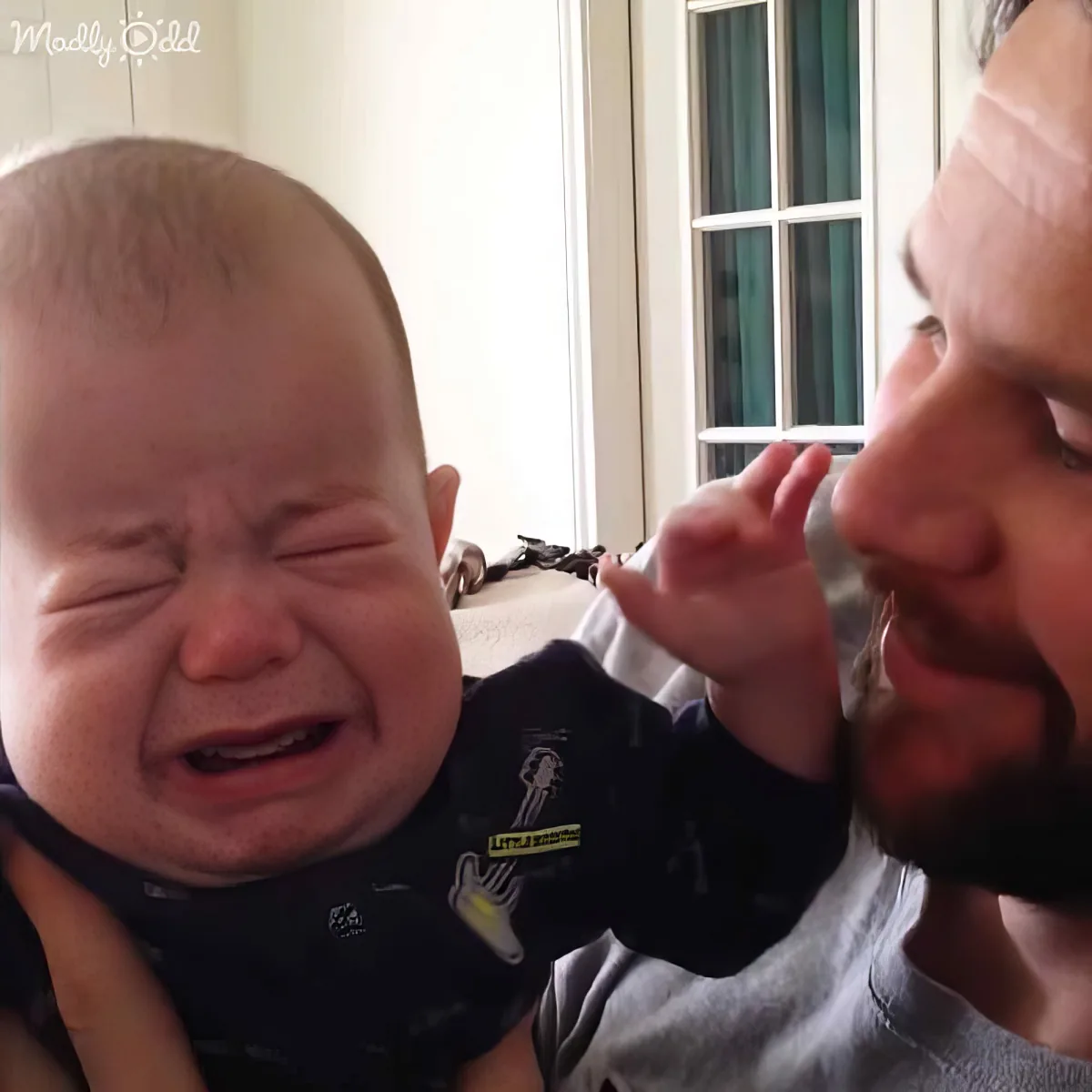 A baby in a black outfit is crying while being held by a person with a beard.